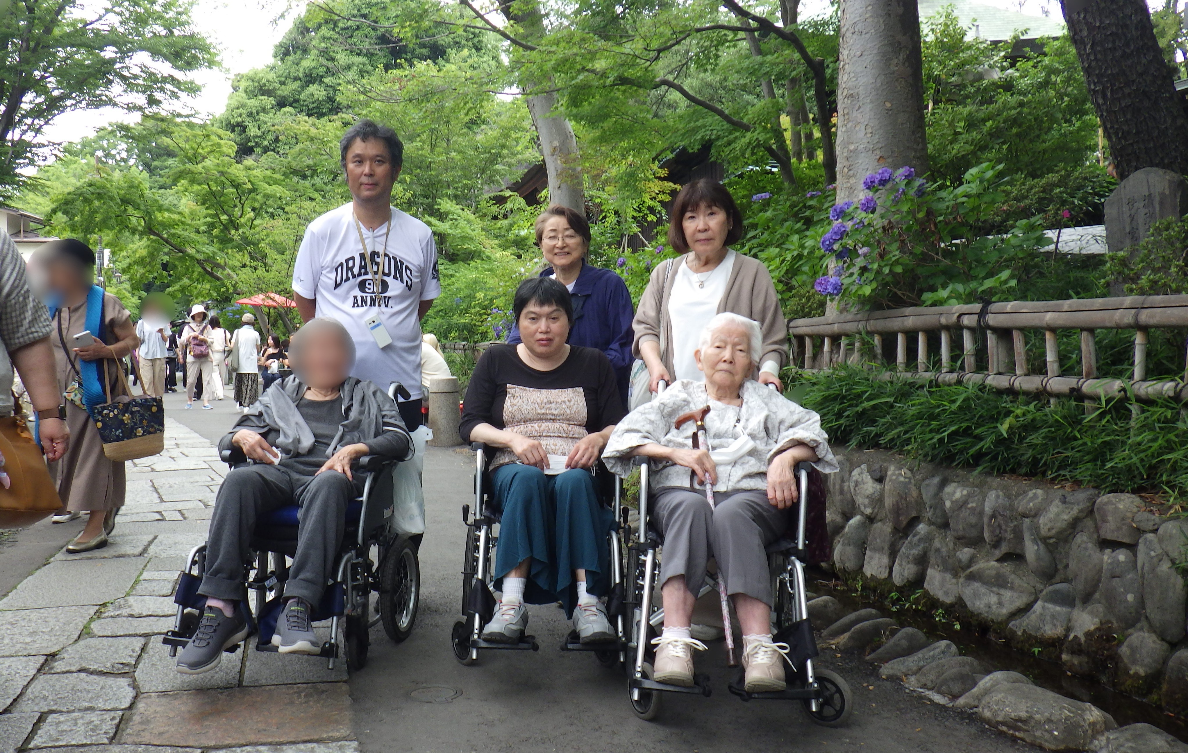 介護施設の利用者と職員が屋外散策・レクリエーション活動を行う様子。車椅子ユーザーも含まれている。